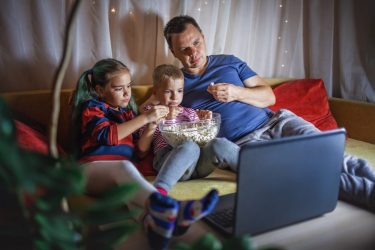 Family movie night. Father with kids watching movie with popcorn by laptop, indoor lifestyle